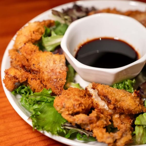 Crispy fried soft-shell crab tempura with a side of dark dipping sauce on a bed of greens.