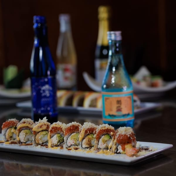 Sushi rolls with avocado, sauce, and crunchy topping, with sake bottles in the background.