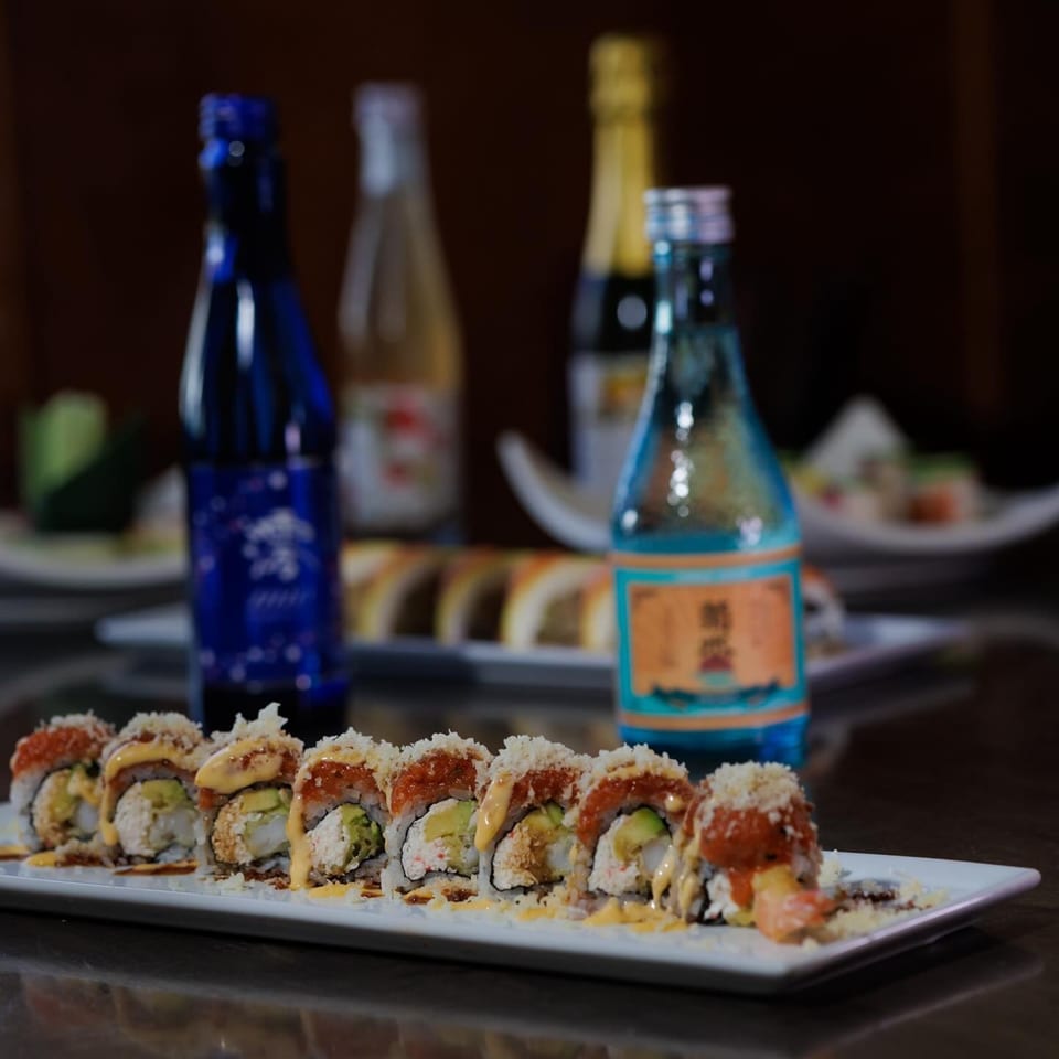 Sushi rolls with avocado, sauce, and crunchy topping, with sake bottles in the background.