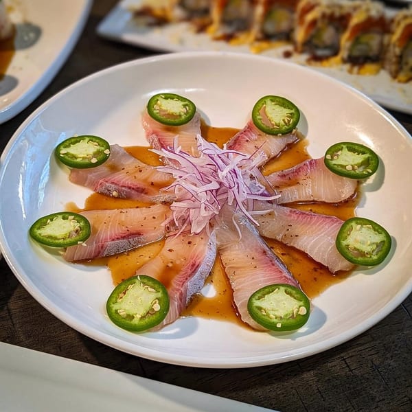 Sake Sushi's yellowtail sashimi with jalapeño slices, red onions, and ponzu sauce on a white plate.